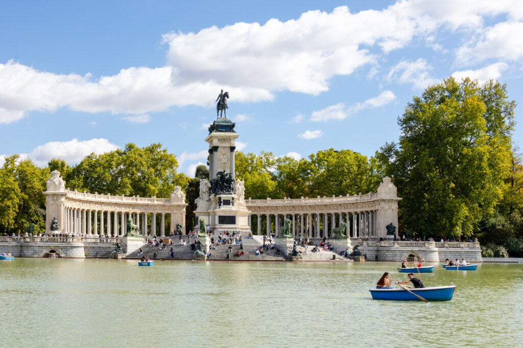 people rowing boats on the pond in Retiro park madrid with people sitting on the steps of the curved, columned memorial