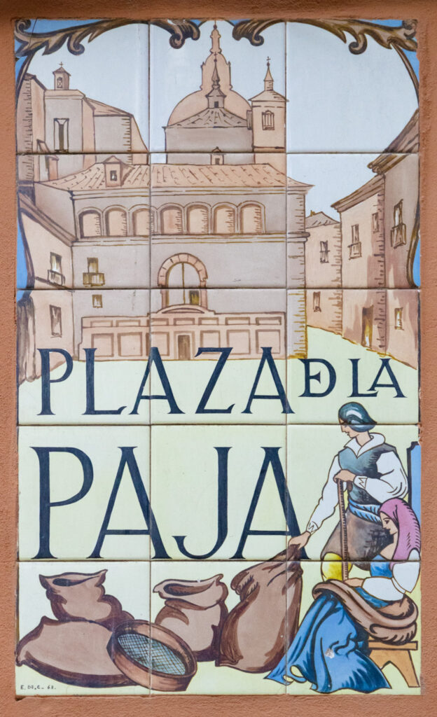 painted tile sign for plaza de la papa madrid that shows a man and woman with bags of straw in the plaza with church behind