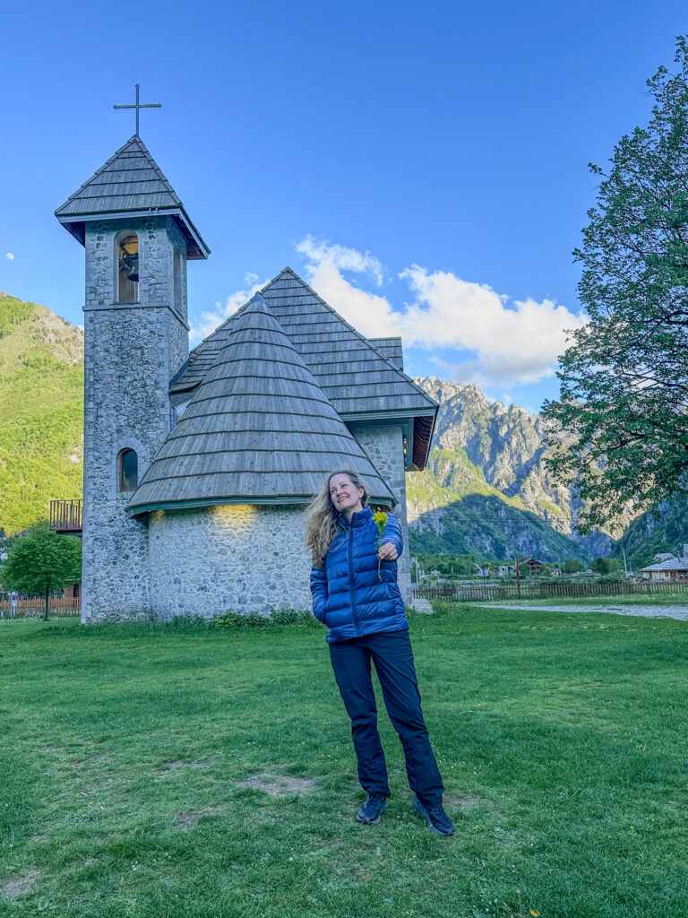 author holding out a flower in front of Kisha e thethit Theth church